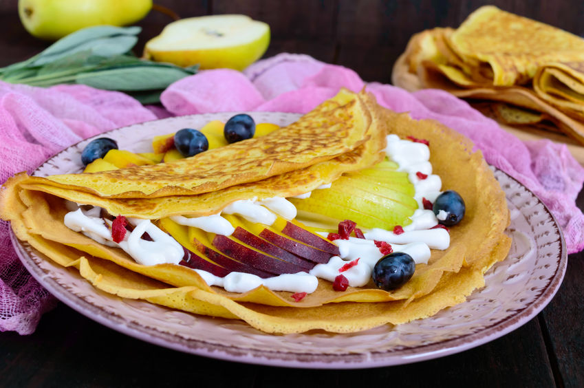 Golden crepes with pieces of peach, pear, cream and grapes on a ceramic plate on a dark background.